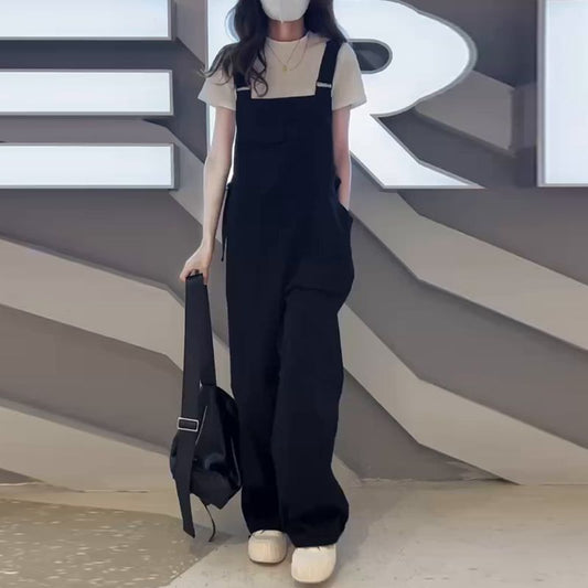 Person wearing black overalls and holding a black bag against a geometric-patterned wall.