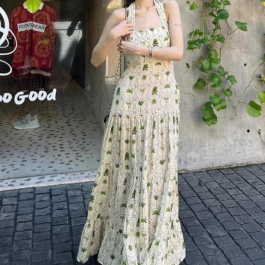 Woman wearing a long floral dress standing on a sidewalk with a store and plant in the background.